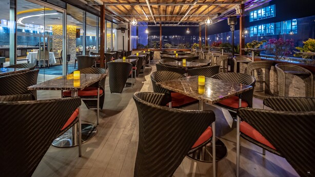 An indoor dining area with wicker chairs and tables, wooden ceiling beams, and a view of the cityscape through large windows. - Sonesta Hotel Arequipa (Arequipa)