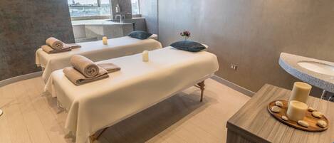 A spa room with two massage tables, a wooden vanity with candles, and a view of the city through a large window.
