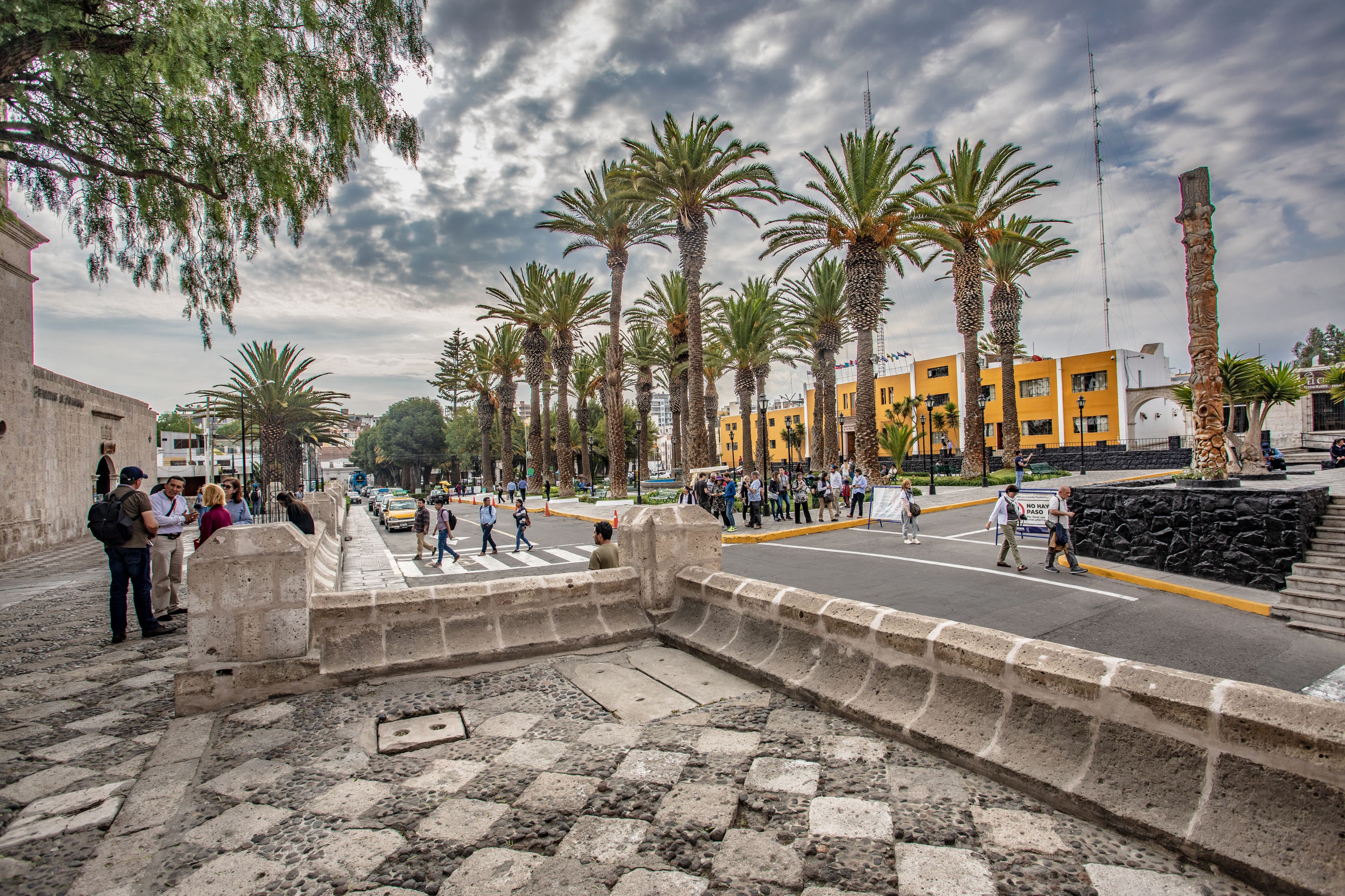 Una calle con palmeras, un edificio amarillo y un muro de piedra.