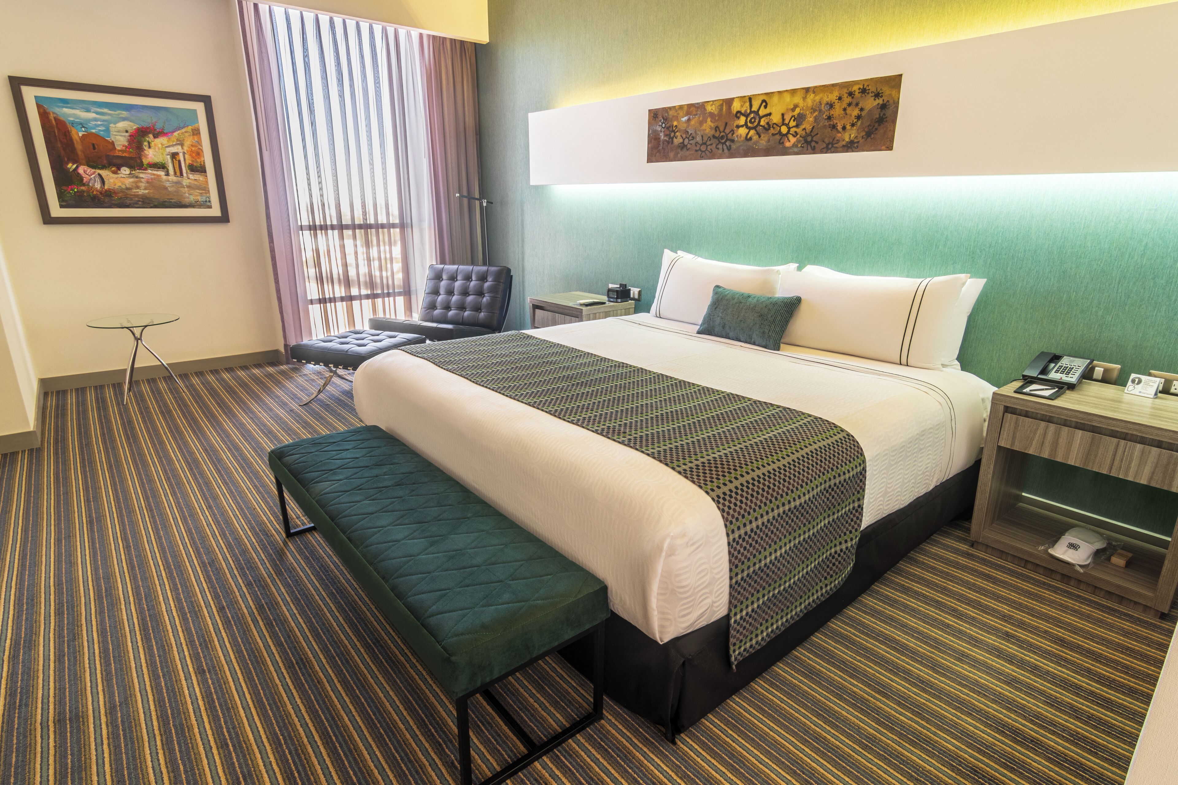 A hotel room with a large bed, a bench, a chair, a small table, and a nightstand with a phone.