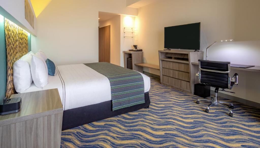 Une chambre d’hôtel comprenant un lit, un bureau, une chaise, une télévision, une lampe et un tapis à motifs.