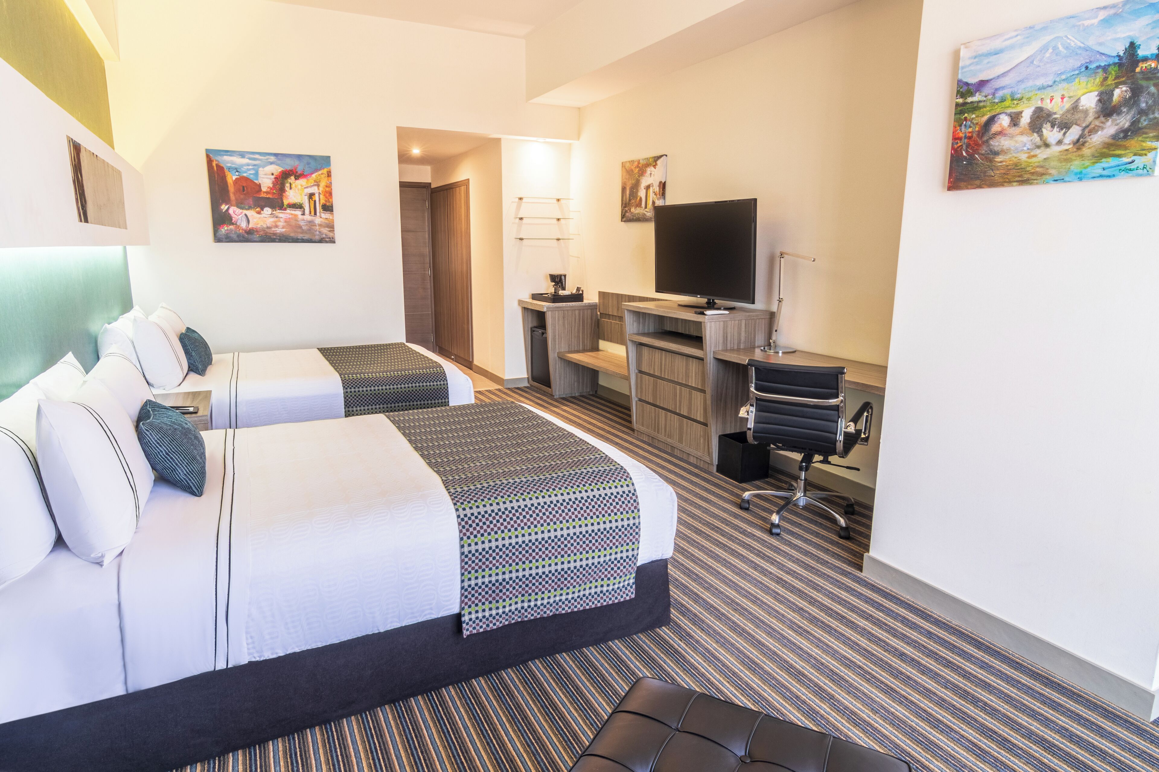 A hotel room with two beds, a desk, a television, and artwork on the walls.