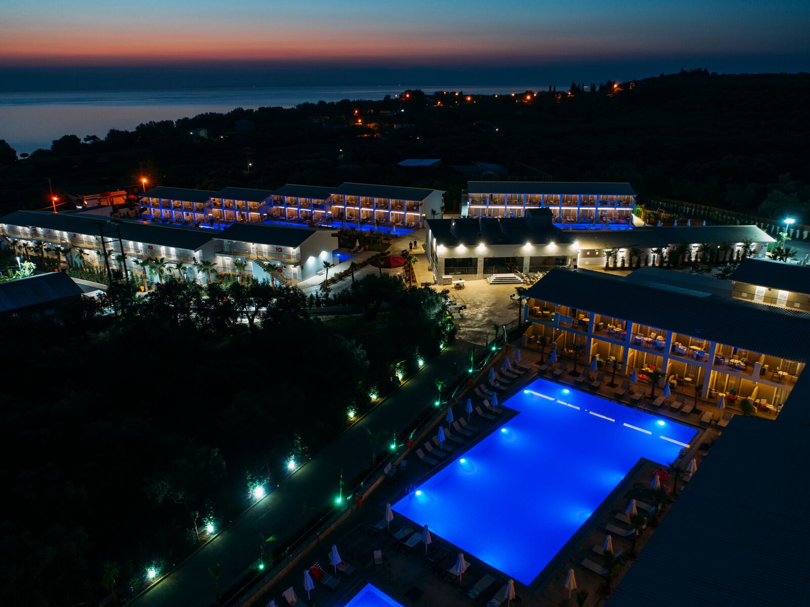 A resort with multiple buildings, a large pool, and a view of the sea at dusk.