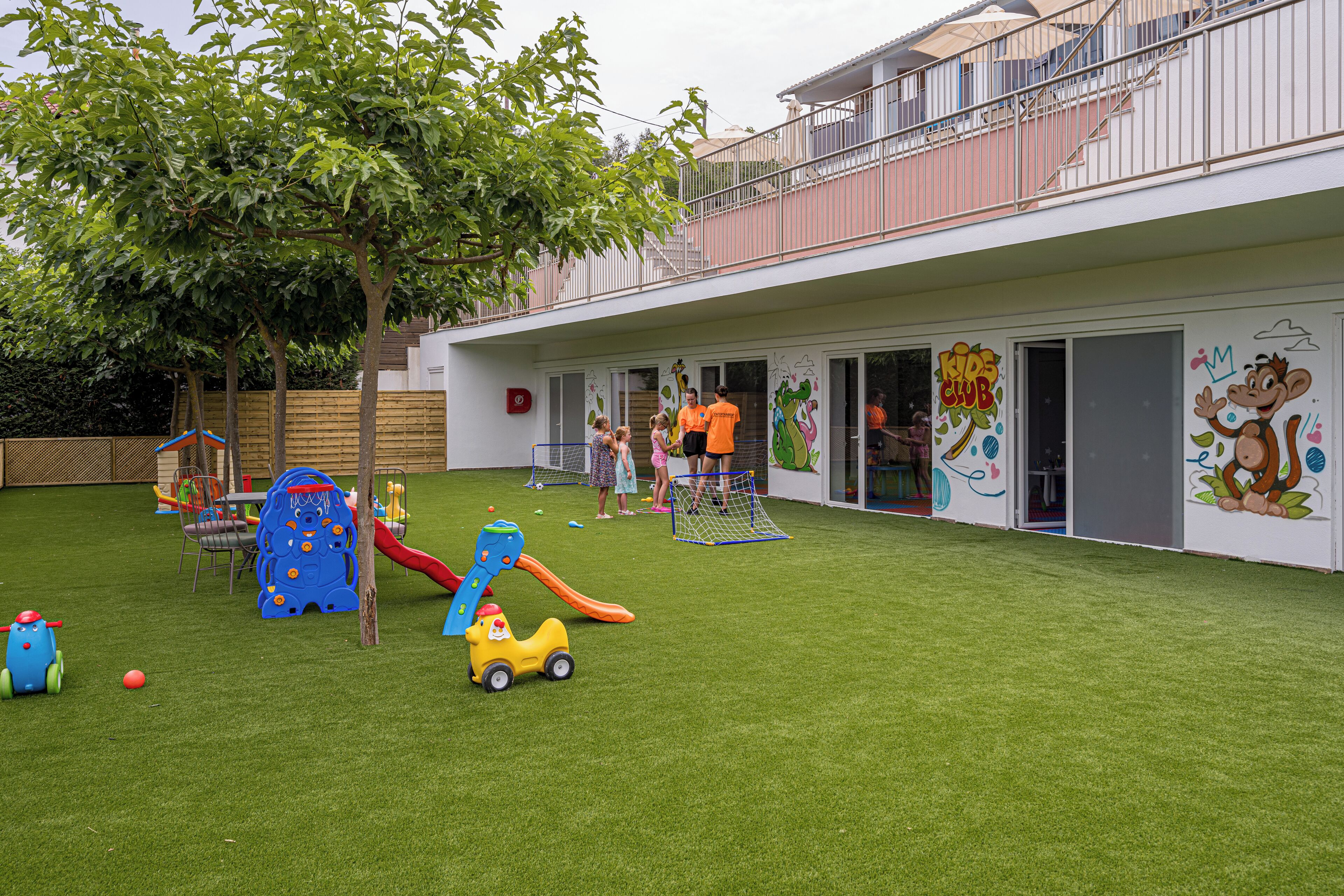 Zona de juegos infantiles con tobogán, pared de escalar y varios juguetes sobre césped artificial.