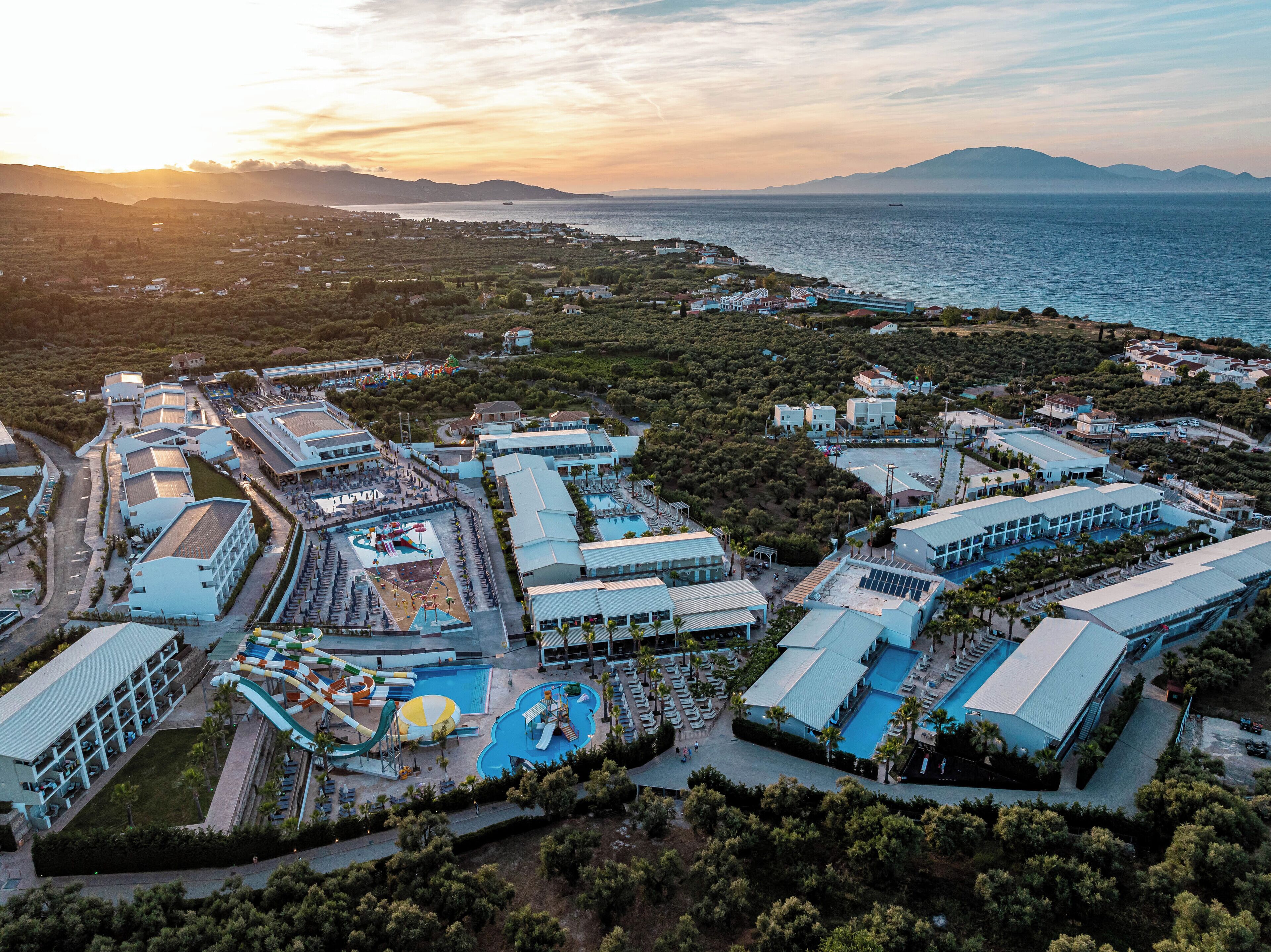 An aerial view of a resort with multiple buildings, a water park, and a large body of water in the background.