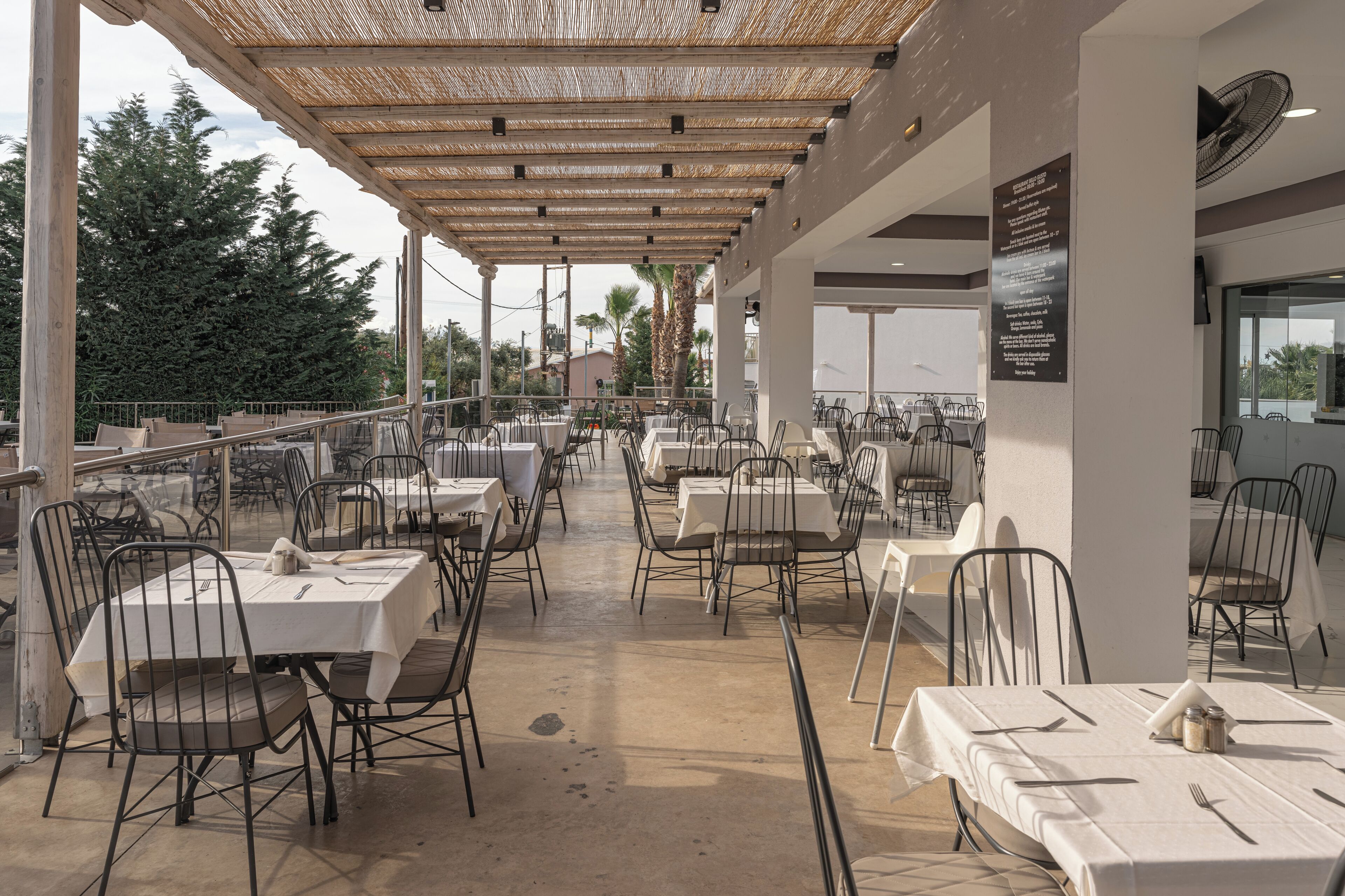 An outdoor dining area with tables and chairs, covered by a woven ceiling, and a view of trees and buildings in the background.