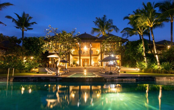 A large, two-story house with a swimming pool in front, surrounded by palm trees and well-lit outdoor areas.