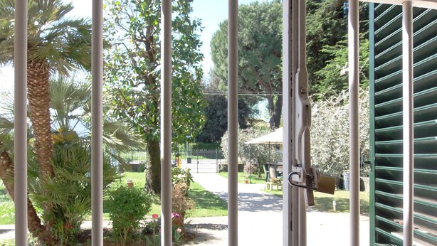Une vue à travers une grille métallique fermée par un cadenas, donnant sur un espace pavé agrémenté de verdure et où l’on aperçoit une clôture au fond.