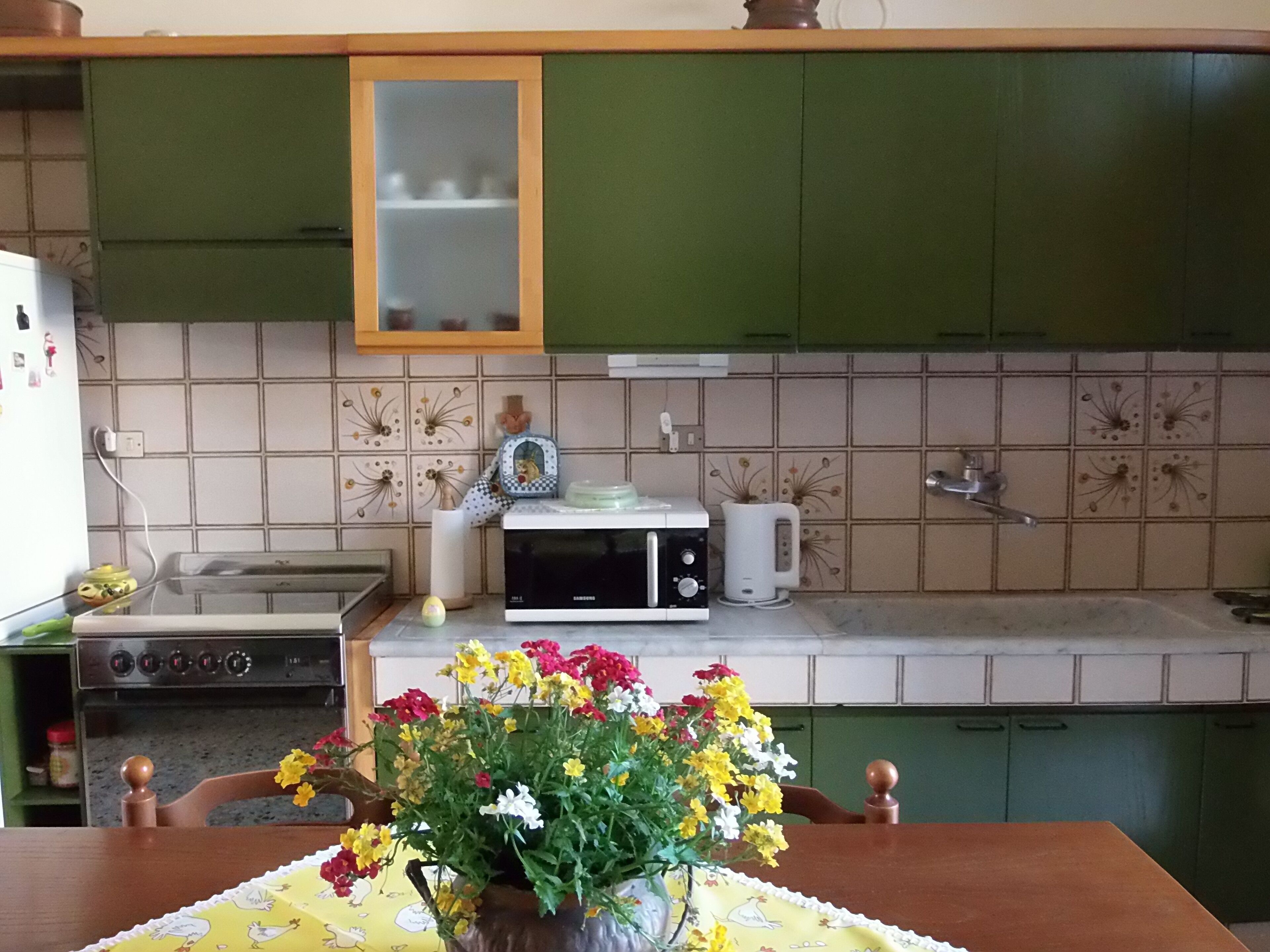 Eine Küche mit grünen Schränken, einer Mikrowelle, einem Wasserkocher und einem Herd. Auf dem Tisch steht eine Vase mit Blumen.