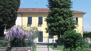 Un bâtiment jaune avec un toit recouvert de tuiles rouges, une porte en métal verte et un arbre aux fleurs violettes devant.