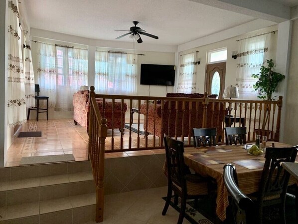 A dining area with a wooden table and chairs, a television mounted on the wall, and a staircase leading to another level. - The Seclusive Mansion, Copthall (Roseau)
