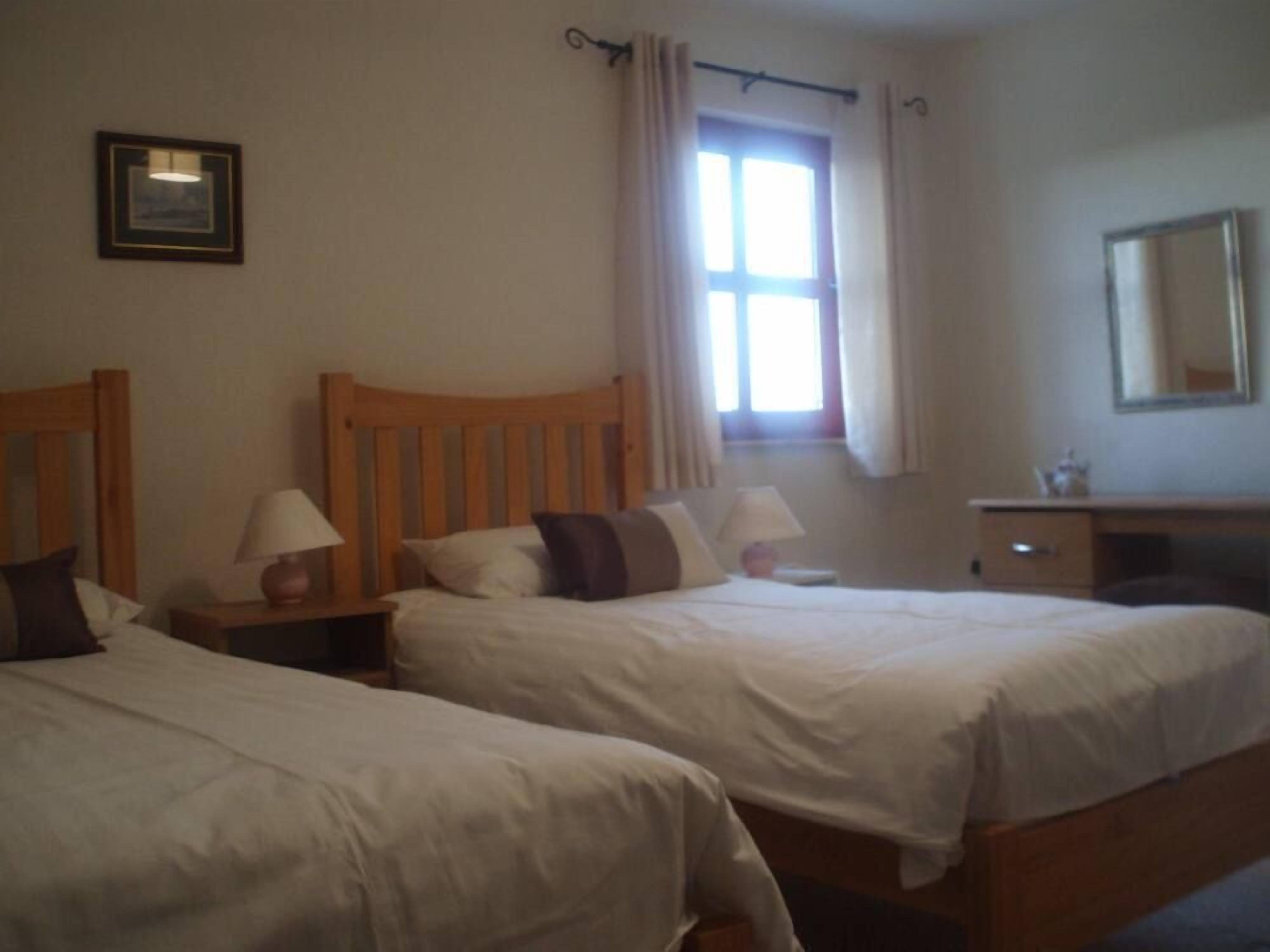 Une chambre d’hôtel avec deux lits simples, une tête de lit en bois, une table de chevet avec une lampe, une commode et une fenêtre avec des rideaux.