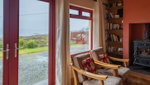 Une pièce chaleureuse avec une grande fenêtre, une chaise en bois dotée d’un coussin, une bibliothèque et un poêle à bois.