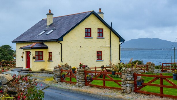 Une maison à deux étages avec une porte et des fenêtres rouges, une clôture en pierre et une vue sur la mer.