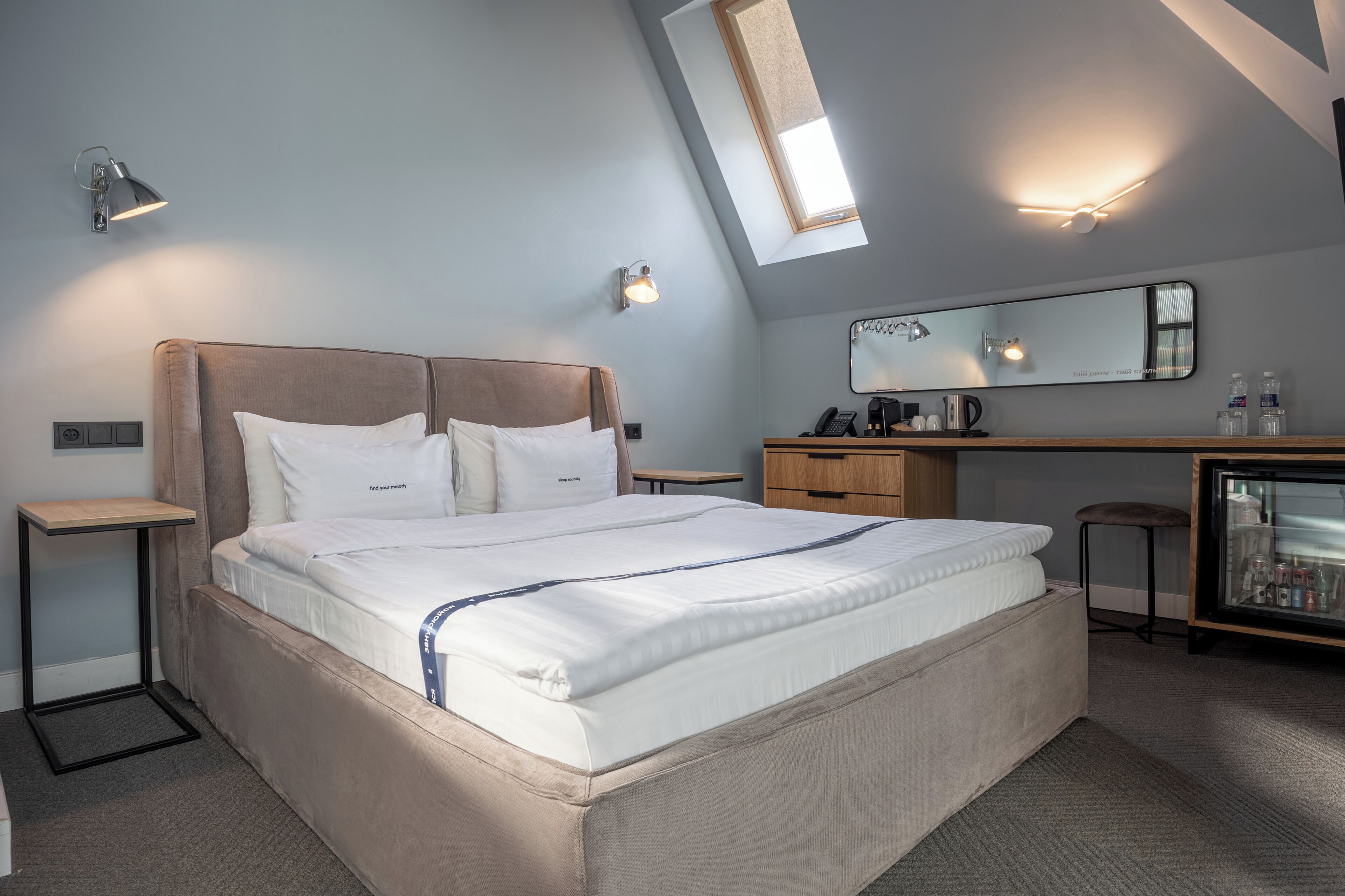 A modern hotel room with a large bed, bedside tables, a desk, a mirror, and a minibar.