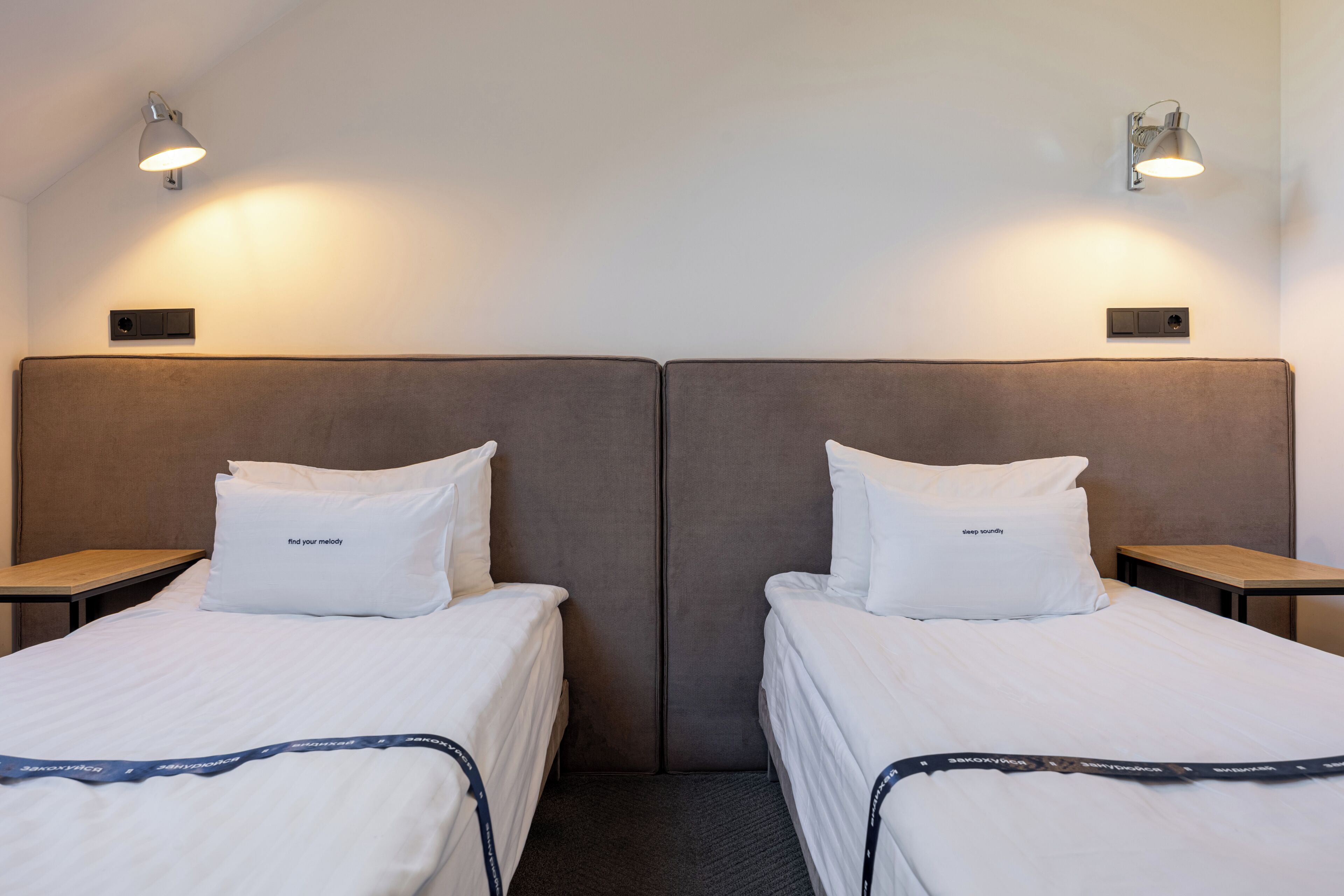A hotel room with two single beds, each with a pillow and a bedside table.