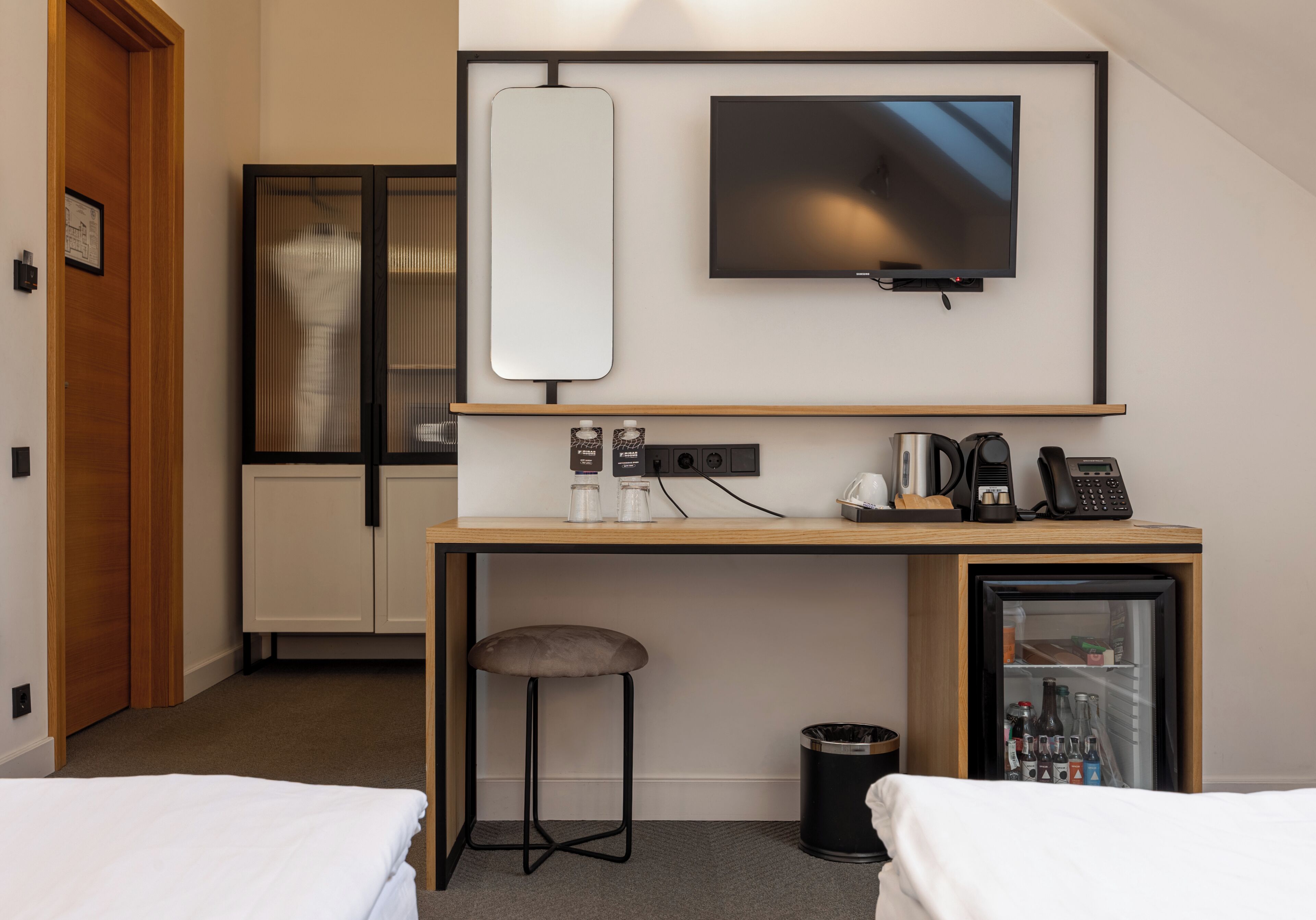 Ett hotellrum med en säng, ett skrivbord med en tv, ett litet kylskåp, en papperskorg och en liten pall.
