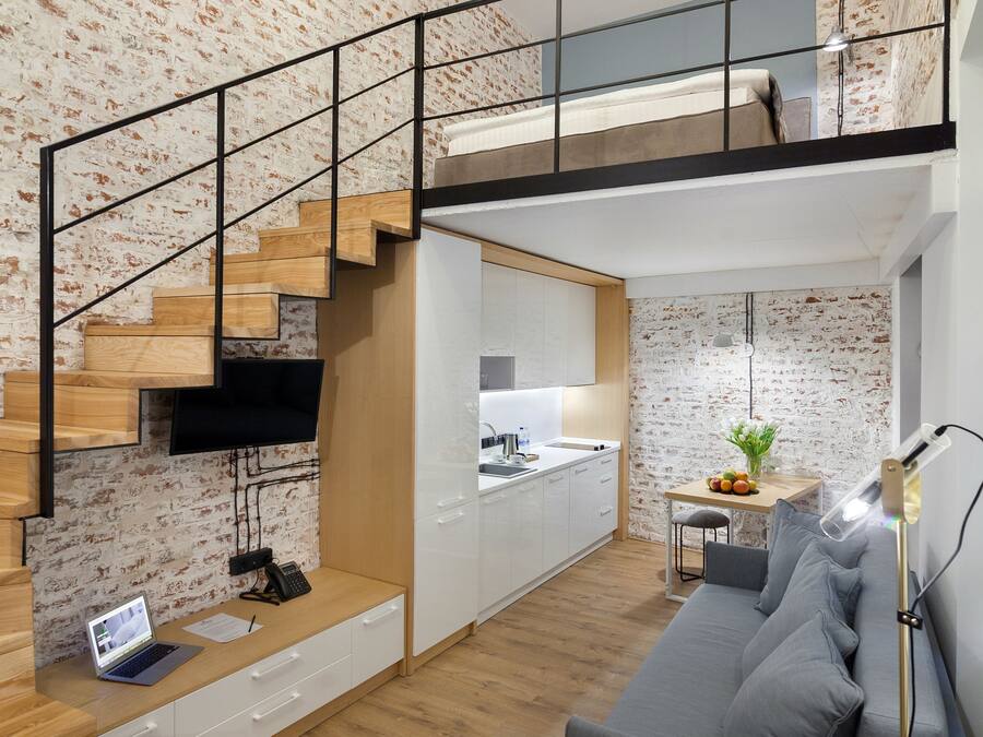Um apartamento moderno estilo loft, paredes de tijolo aparente, escada de madeira, mezanino, cozinha e área de estar com sofá e escrivaninha.