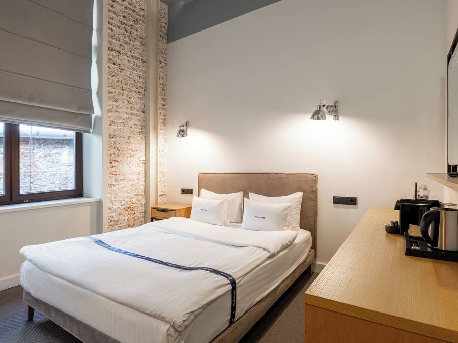 A hotel room with a bed, a desk with a kettle, a brick wall, and a window.