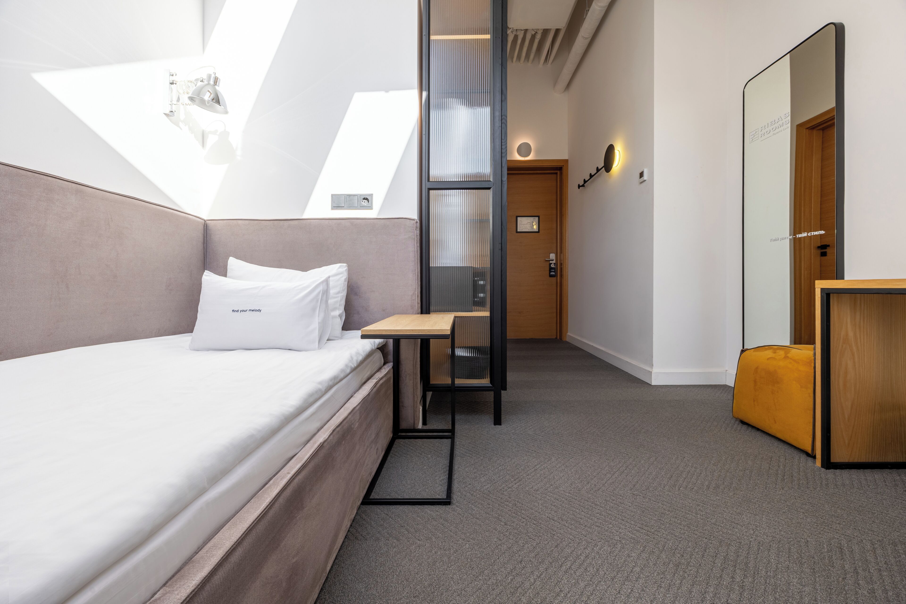 A modern hotel room with a bed, a small table, a mirror, and a door.