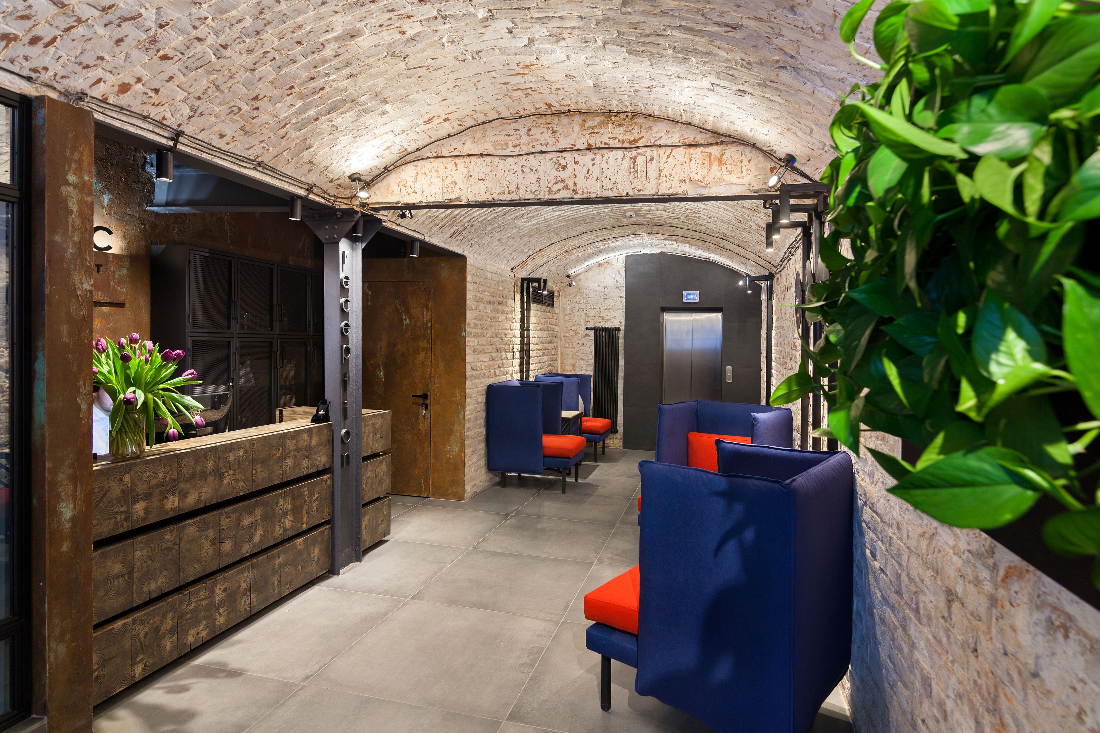 A modern lobby with a brick ceiling, wooden reception desk, blue seating, and a plant.