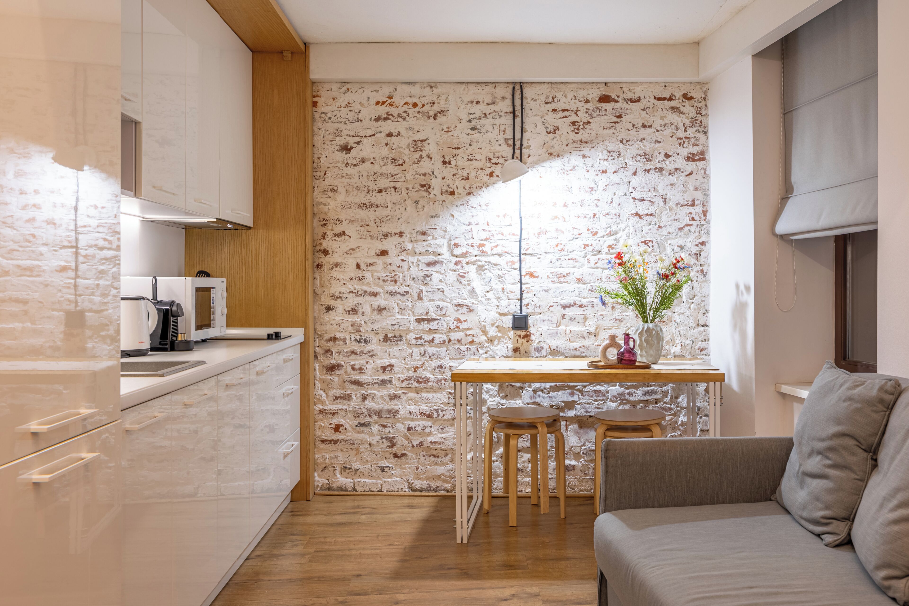 Une cuisine compacte avec des meubles blancs, un plan de travail en bois, un mur en briques, une petite table à manger avec deux tabourets et un canapé.