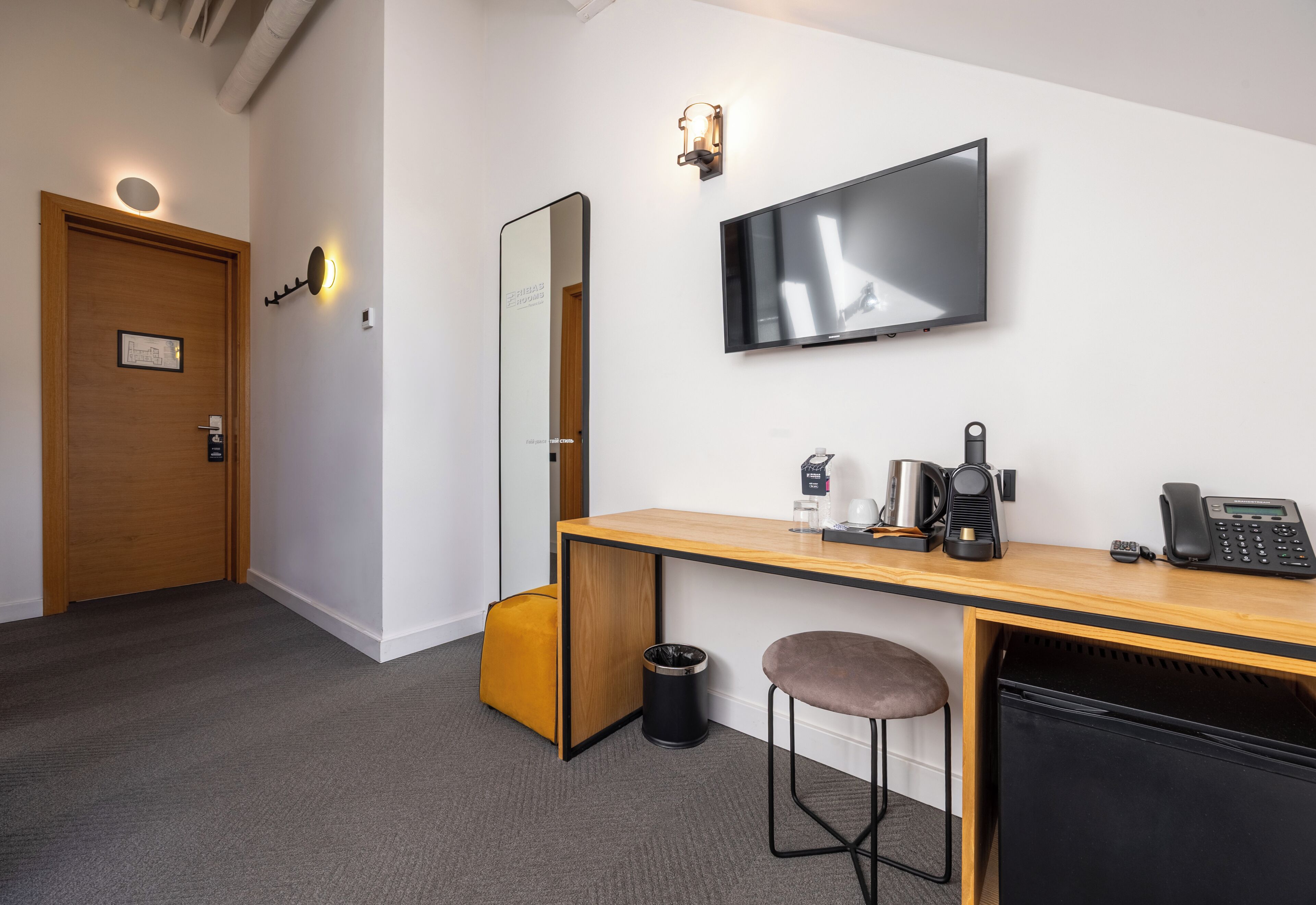 Habitación de hotel con escritorio de madera, televisor de pantalla plana, máquina de café, teléfono, cubo de basura, taburete acolchado y puerta con lámpara.
