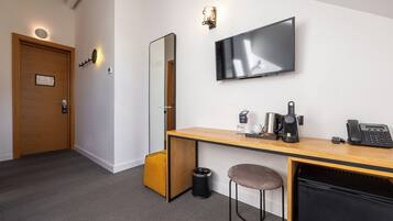 Habitación de hotel con escritorio de madera, televisor de pantalla plana, máquina de café, teléfono, cubo de basura, taburete acolchado y puerta con lámpara.