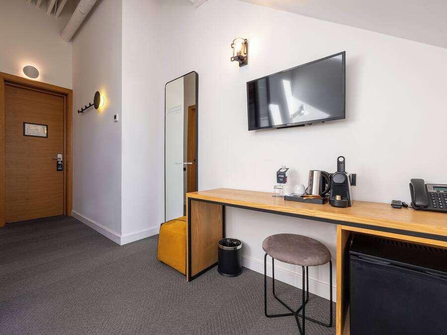 Habitación de hotel con escritorio de madera, televisor de pantalla plana, máquina de café, teléfono, cubo de basura, taburete acolchado y puerta con lámpara.