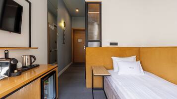 A hotel room with a bed, a desk with a kettle, a television, and a refrigerator.