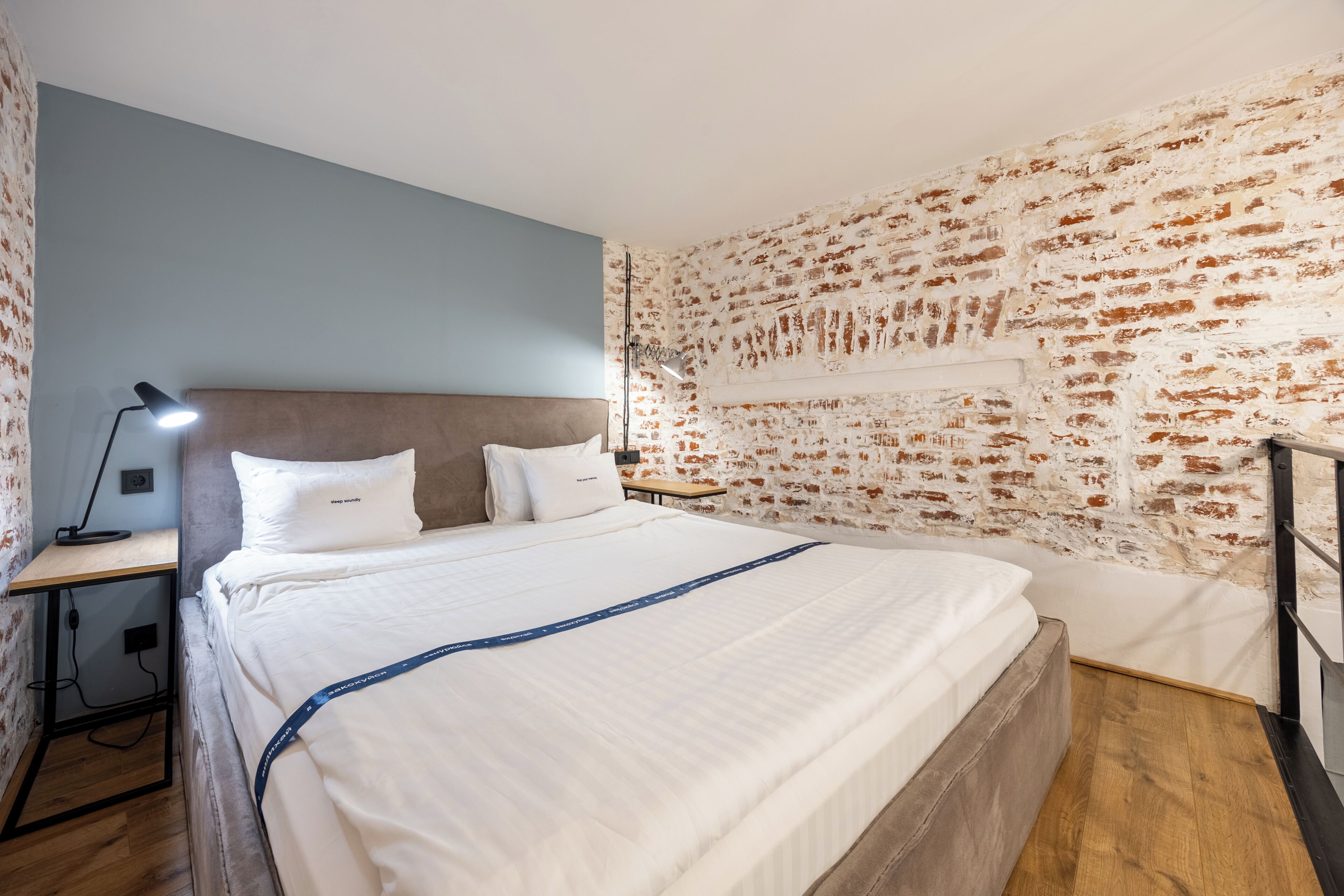 Quarto com parede de tijolos aparentes, cama com roupa de cama branca, mesa de cabeceira com abajur e piso de madeira.