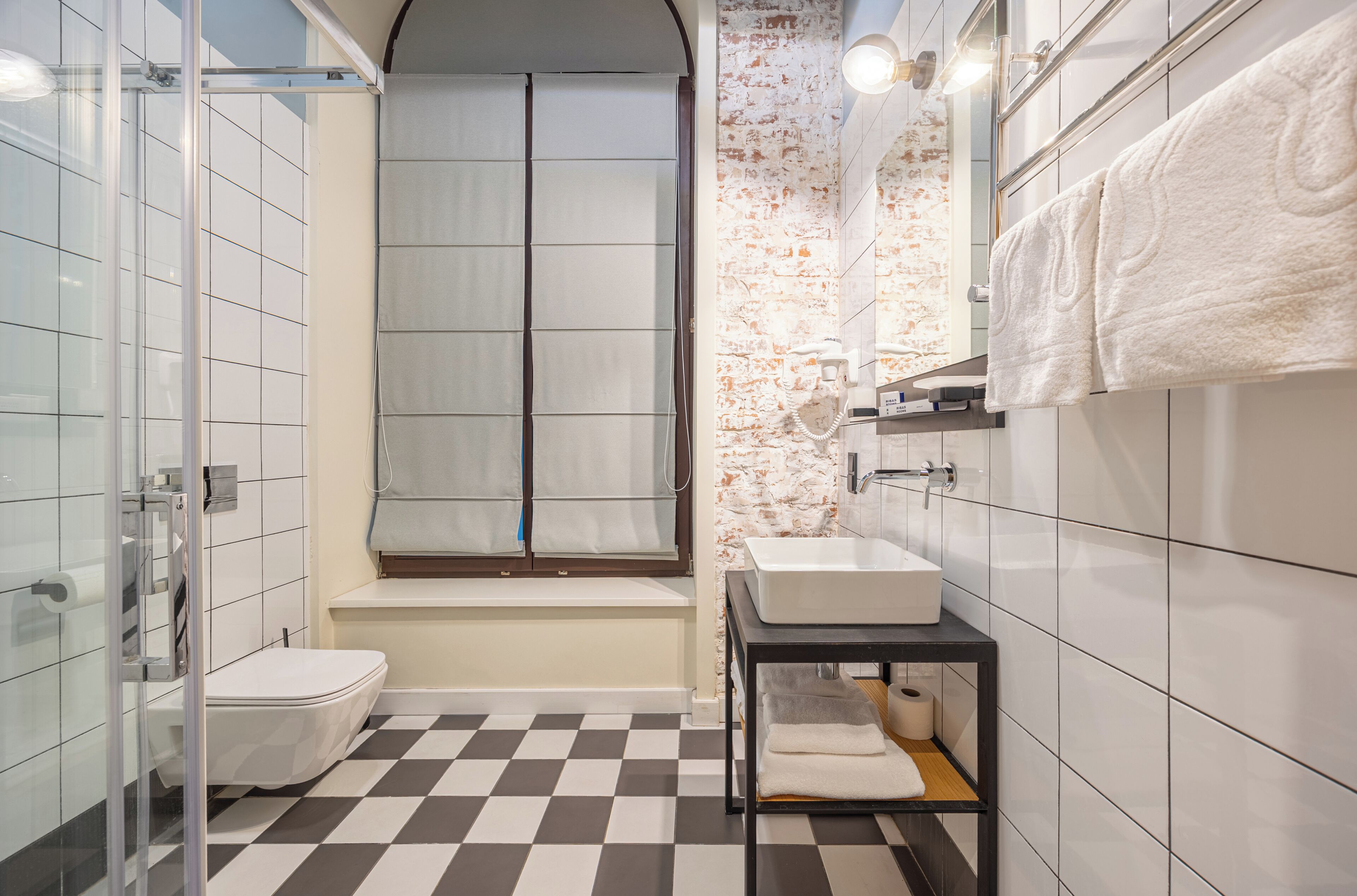 Un baño moderno con piso a cuadros, cabina de ducha de vidrio, lavamanos blanco y ventana con vistas.