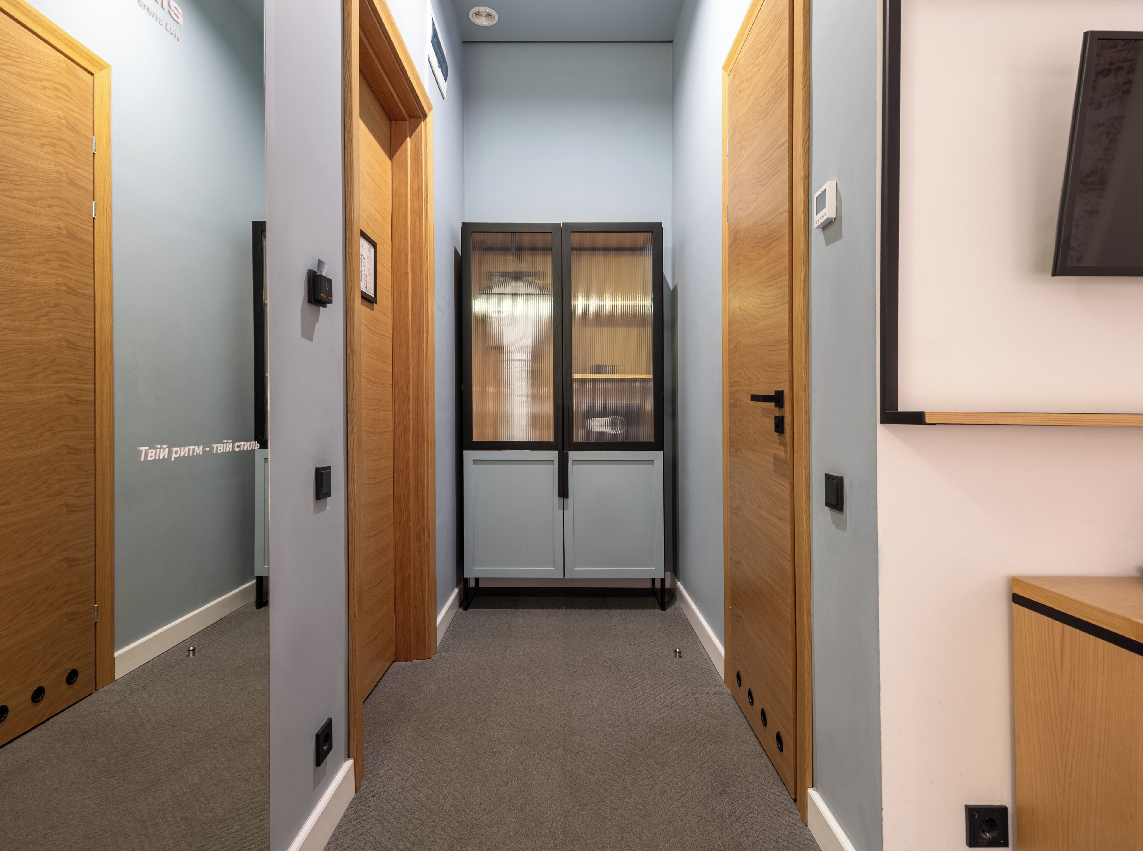 Un couloir étroit avec des portes en bois, une fenêtre avec des stores et un interrupteur sur le mur.