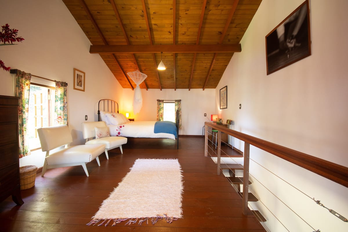 A cozy bedroom with a wooden ceiling, a bed with white bedding, a white sofa, and a large window with floral curtains.