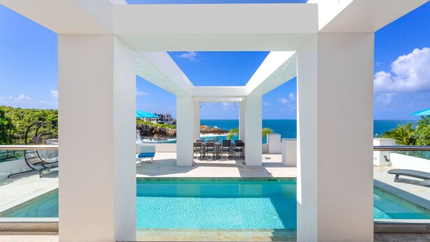 Zona de piscina con vista al océano, rodeada de pilares blancos y un cielo azul despejado.