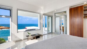Un dormitorio moderno con grandes ventanales con vistas al mar, un armario de madera y una mesita de noche con un florero.