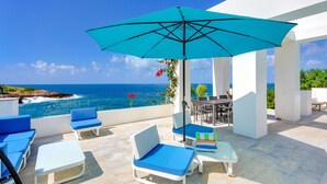 Una terraza con sillones azules, una mesa y un paraguas turquesa con vista al océano.
