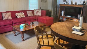 Una sala de estar acogedora con un sofá rojo, una mesa de centro de madera y una mesa de comedor con sillas. Un ventilador de techo y un televisor están montados en la pared.