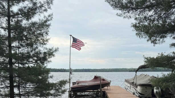 Terraza junto al lago con un brasero, sillas Adirondack y un muelle con un bote.
