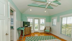A bedroom with a bed, dresser, television, and a view of the outdoors through large windows. - Large home with private guest crofter across from the beach access, wrap around porches (Bald Head Island)