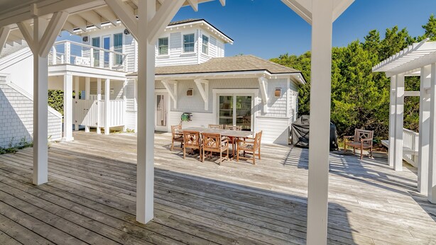 A wooden deck with a dining table and chairs, surrounded by white columns and a covered area. - Large home with private guest crofter across from the beach access, wrap around porches (Bald Head Island)