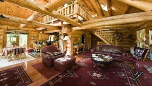 Interior de una cabaña de troncos con techo de madera, comedor, sala de estar con muebles de cuero y una escalera que conduce a un nivel superior.