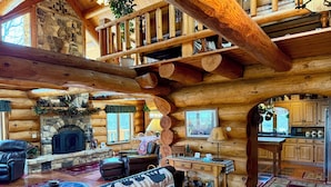 Interior de una cabaña de troncos con chimenea de piedra, mobiliario de cuero y zona de cocina.
