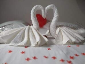 Dos toallas con forma de cisne, con un corazón en el centro, sobre una cama con sábana blanca y motivos de hojas de arce rojas.
