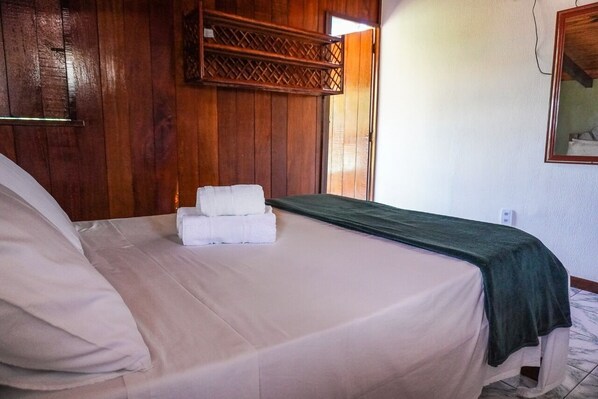 Una cama bien hecha con una manta verde y dos toallas blancas, paredes de paneles de madera y una ventana pequeña.