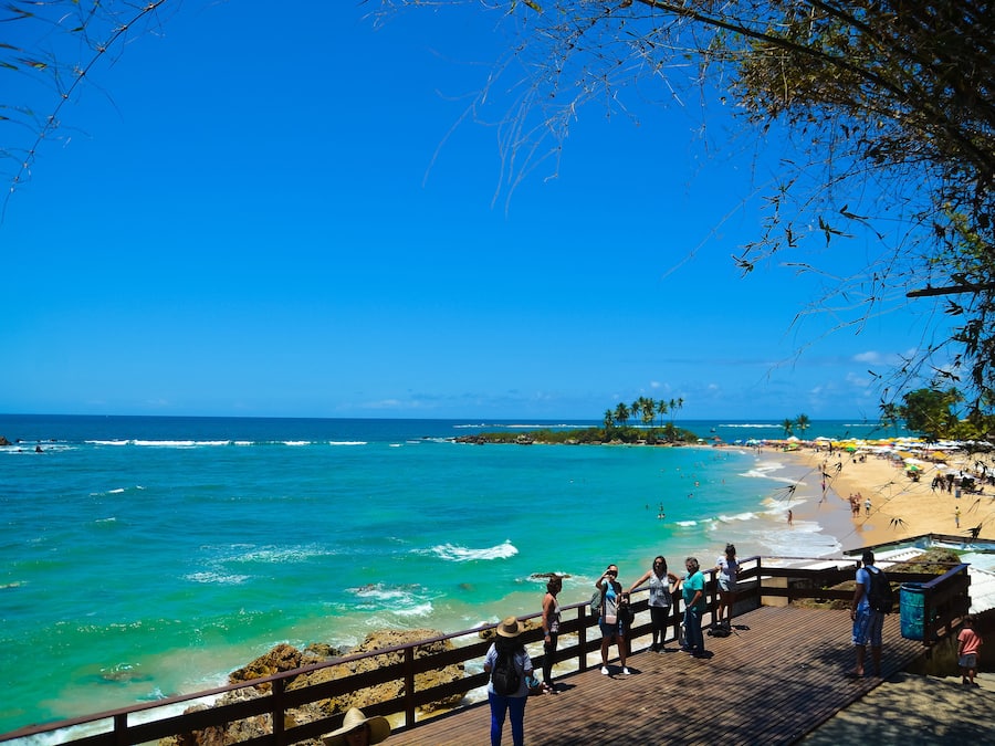 Uma praia com calçadão de madeira, água turquesa cristalina e céu azul transparente.