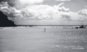 A beach scene with a person paddleboarding, waves, and a cliff in the background. - Pousada da Praça (Cairu)