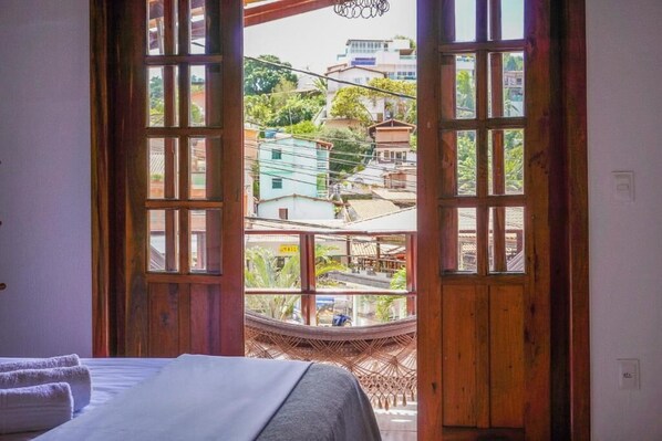 A room with a bed, a view through a wooden-framed window, and a woven chair outside. - Pousada da Praça (Cairu)