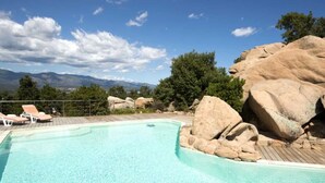 A pool with a wooden deck, surrounded by large rocks and trees, with a view of mountains in the background. - Magnificent air-conditioned villa with private pool, 2 bedrooms 2 bathrooms (4 people) (Porto-Vecchio)