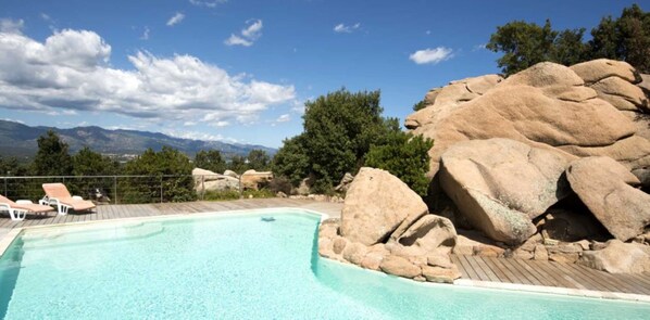 A pool with a wooden deck, surrounded by large rocks and trees, with a view of mountains in the background. - Magnificent air-conditioned villa with private pool, 2 bedrooms 2 bathrooms (4 people) (Porto-Vecchio)